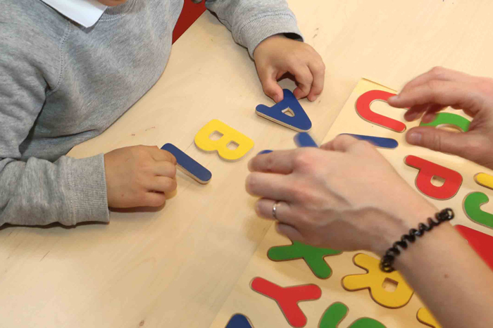 Seules les mains sont visibles, un enfant manipule des lettres de l'alphabet, aidé par une femme